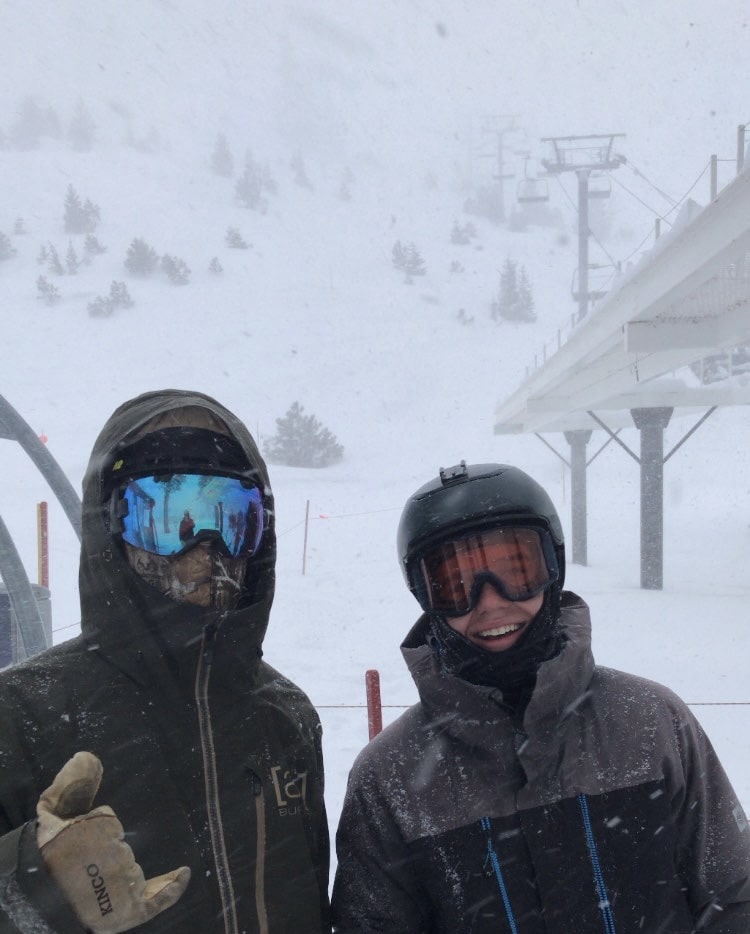 Two riders smile at the camera while snow comes down around them.