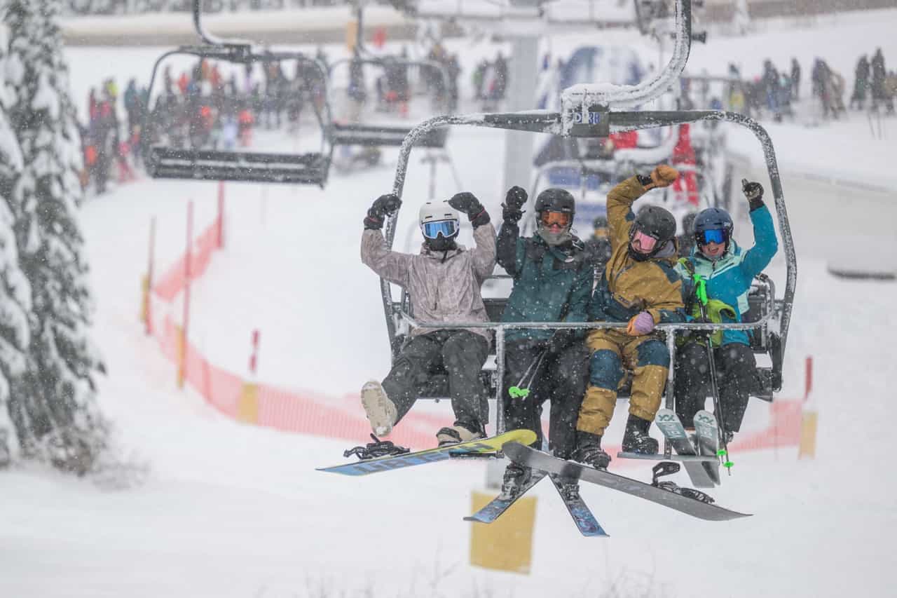 happy skiers and boarders on a chairlift at a snowy big white resort on opening day