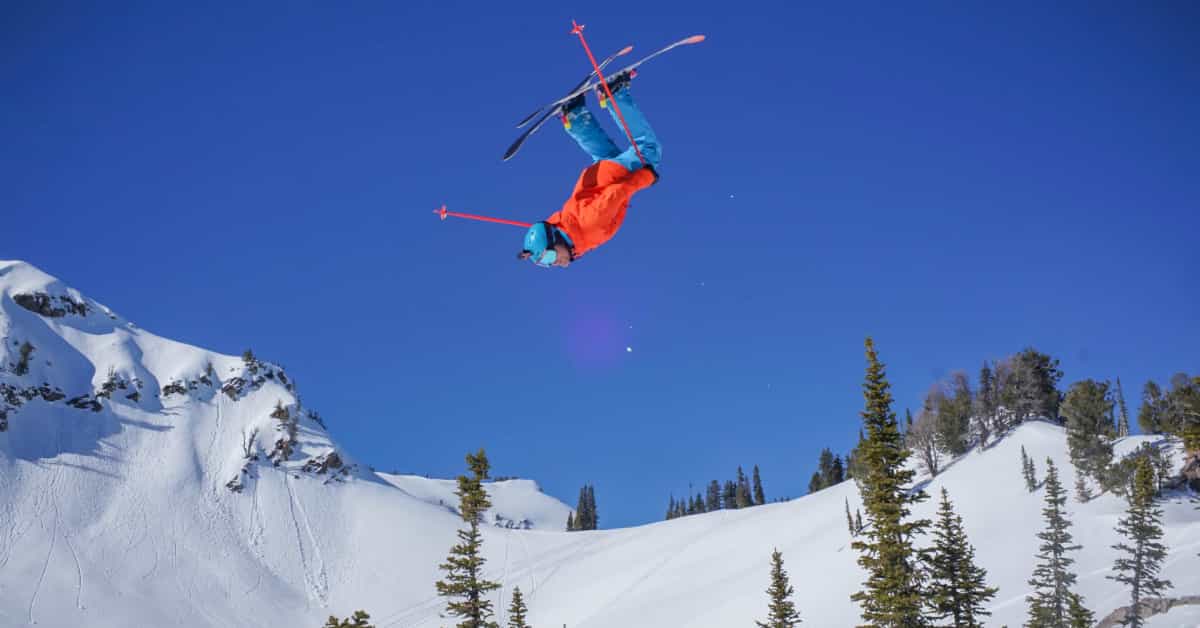 skier owen leeper throwing a huge front flip off a cliff with blue skies and snowy mountains in the background