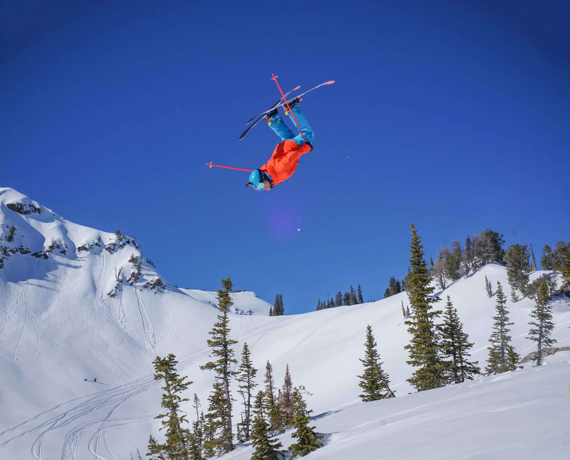 skier owen leeper throwing a huge front flip off a cliff with blue skies and snowy mountains in the background